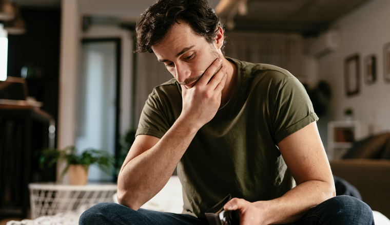 Young man checking empty wallet, no money. Unemployed sad man sitting at home thinking about money.