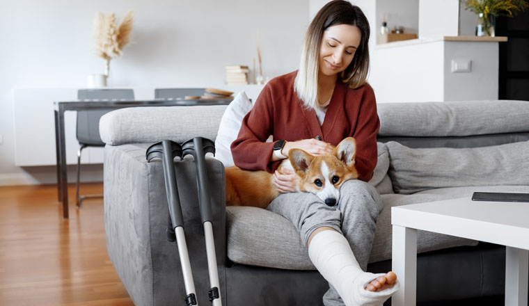 Adult woman in her late twenties on couch at home with crutches and orthopedic plaster caress the dog. Fracture of the leg or foot. Concept of rehabilitation and healing. Orthopedics and Traumatology.
