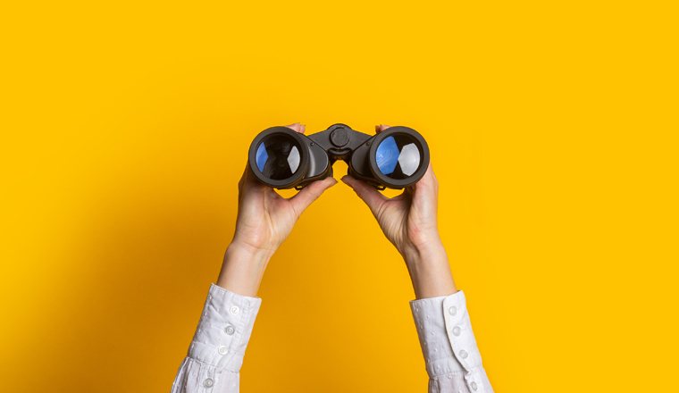 female hands hold black binoculars on a bright yellow background.