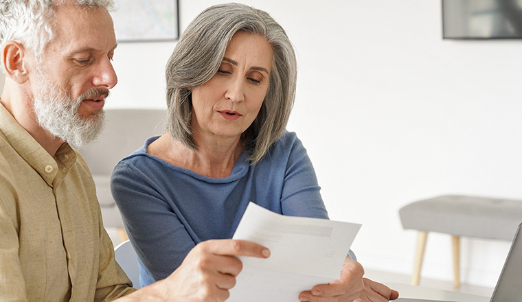 Older mature couple checking bank documents using laptop at home. Senior mid age retired man and woman reading paper bills, calculating pension or taxes, planning retirement finances, doing paperwork.