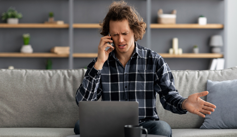 Discontented Man Talking On Phone Using Laptop Having Problem With Computer Or Internet Connection Sitting At Home. Displeased Customer Calling To Hotline Service. Selective Focus