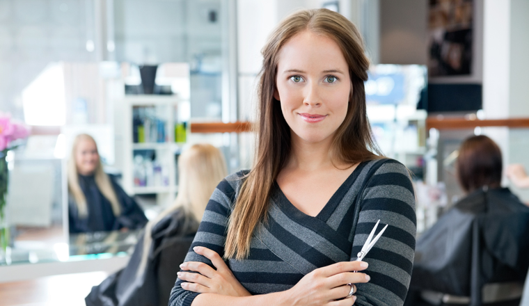 Portrait of a confident female hairdresser standing hands folded with people in background