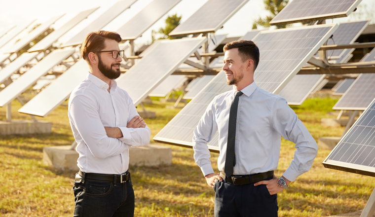 Happy male business partners discussing energy development project while standing near photovoltaic panels on green solar farm