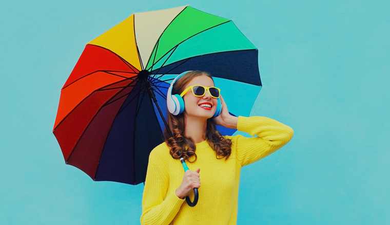 Autumn portrait of happy cheerful smiling young woman with colorful umbrella listening to music in headphones wearing an yellow knitted sweater on blue background