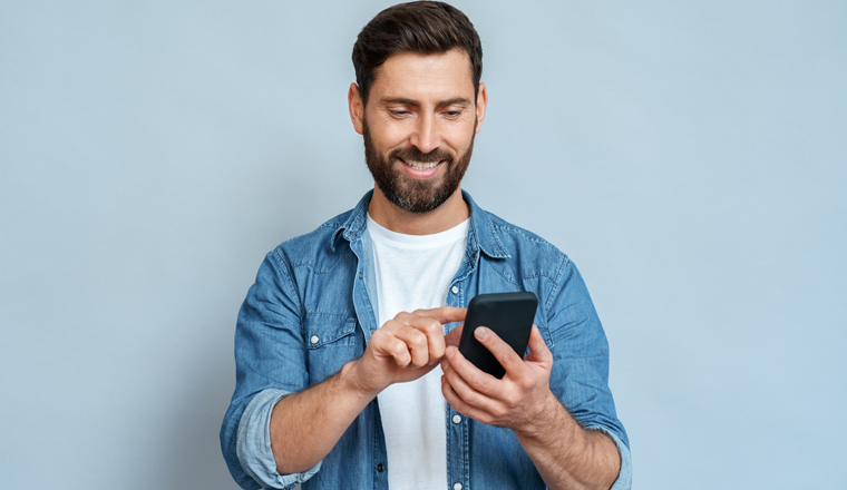 Smiling caucasian man using smartphone modern device looking at mobile screen scrolling enjoying communication with friends. Handsome guy standing isolated on blue background studio copy space