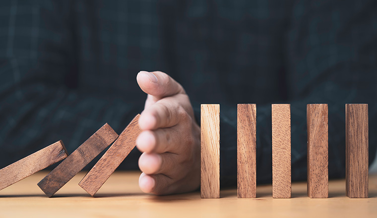 ฺBusinessman uses his hand to stop a falling wooden block to a standing wooden block domino , It is a symbol of protection against damage or stop loss for crisis management concept.