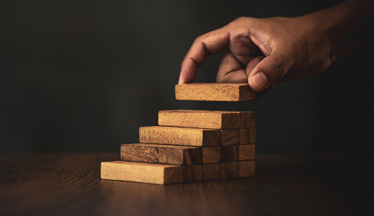 Close-up hand is placing wood block tower stacked in stair step with caution to prevent collapse or crash concepts of financial risk management and strategic planning.