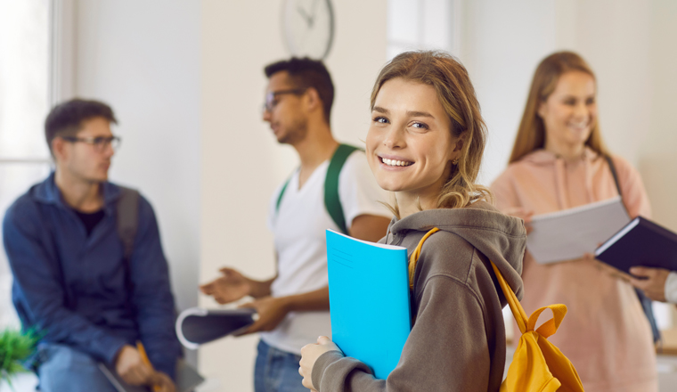 Happy beautiful female university student with book and backpack. Portrait of pretty young girl with candid cheerful face expression smiling at camera while standing in classroom with her classmates