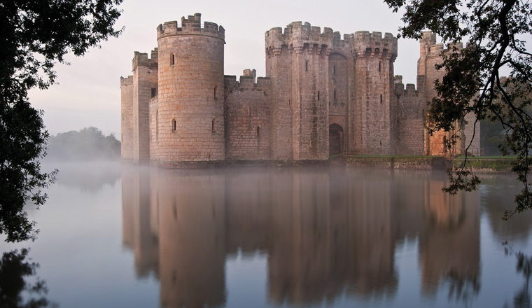 Beautiful medieval castle and moat at sunrise with mist over moat and sunlight behind castle