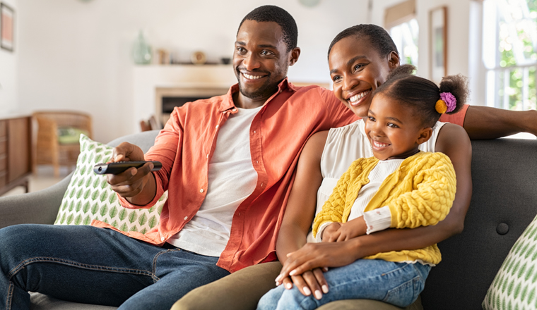 Relaxed african american family watching TV together. Happy mature father changing television channel using remote control with daughter sitting on mother's lap at home. Cheerful black family watching movie together and having fun.