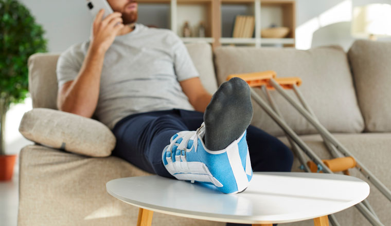Close up of injured man with bandage on leg sit on sofa talk on cellphone with doctor. Unhealthy guy with sling on broken foot suffer from fracture or injury. Rehabilitation at home concept.