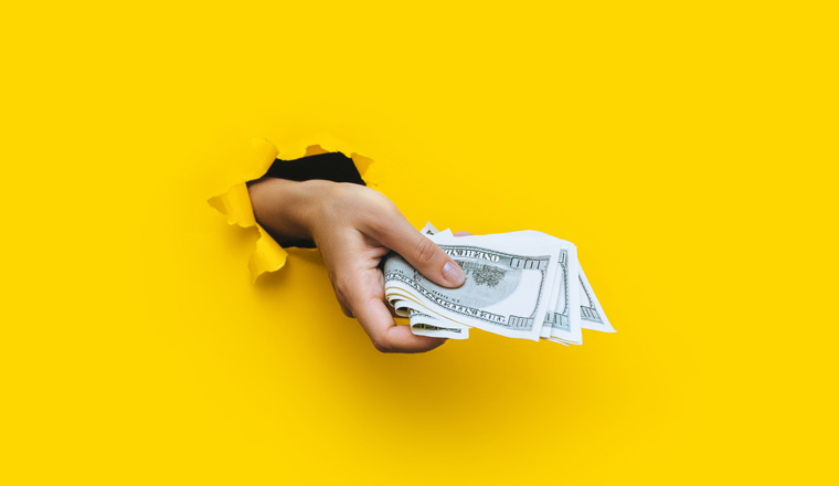 A female hand appears in the hole in torn yellow paper and squeezes several hundred-dollar bills. Concept bribe and benefits, scholarships. Copy space.