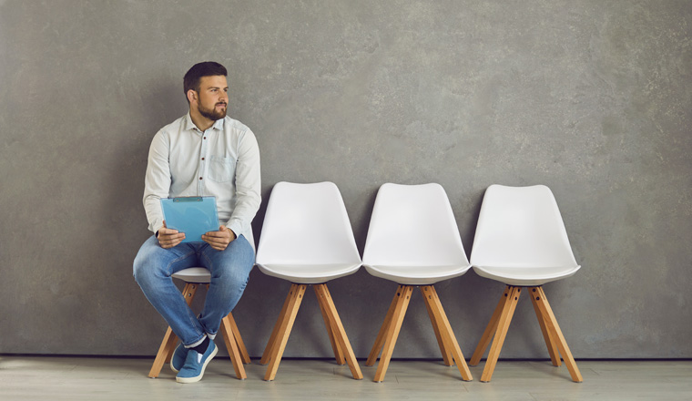 Worried young caucasian male job seeker vacancy candidate holding resume form waiting for interview meeting sitting on chair looking aside. Jobless applicant at recruitment staffing agency