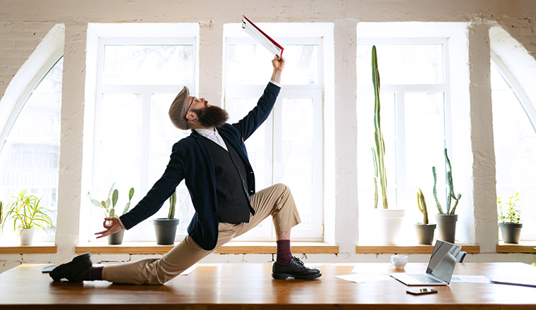 Unusual business process. Young bearded man, office clerk having fun, doing yoga on wooden table in modern office at work time with gadgets. Concept of business, healthy lifestyle, sport, hobby