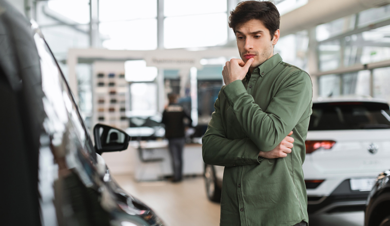 Pensive young guy thinking, making choice, having doubt about buying new car at dealership centre, copy space. Thoughtful millennial Caucasian man selecting auto at showroom store