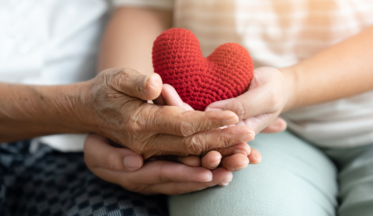 Young and senior woman holding each other hands and red yarn heart shape togetherness concept. Elderly care and protection with love from grandchild. 