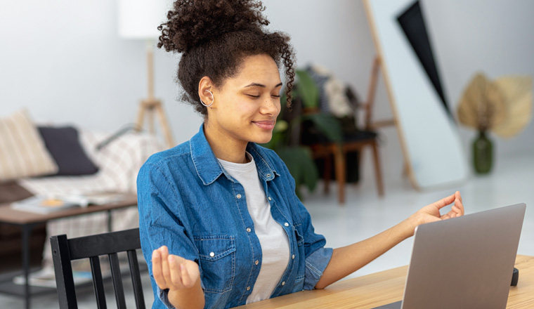 Calm African American young woman sitting at the workplace in the office and meditating taking break avoiding stressful job