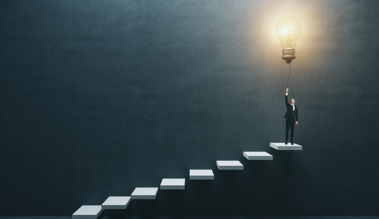 Innovation and idea concept with front view on businessman in suit on the top of white stairway turning on big light bulb on dark concrete background in empty hall