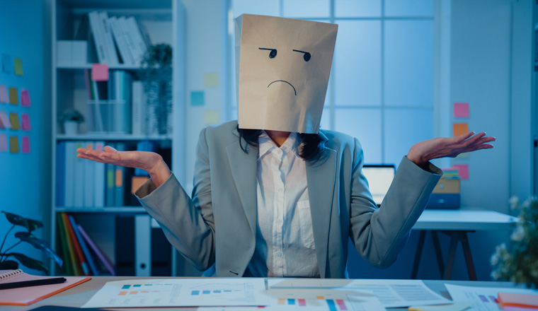 Young Asian businesswoman sit on desk wear draw sad mask paper craft bag feel bad mood with fail work project in office at night. Anonymous lady mad and confuse with overwork, Work mental health.