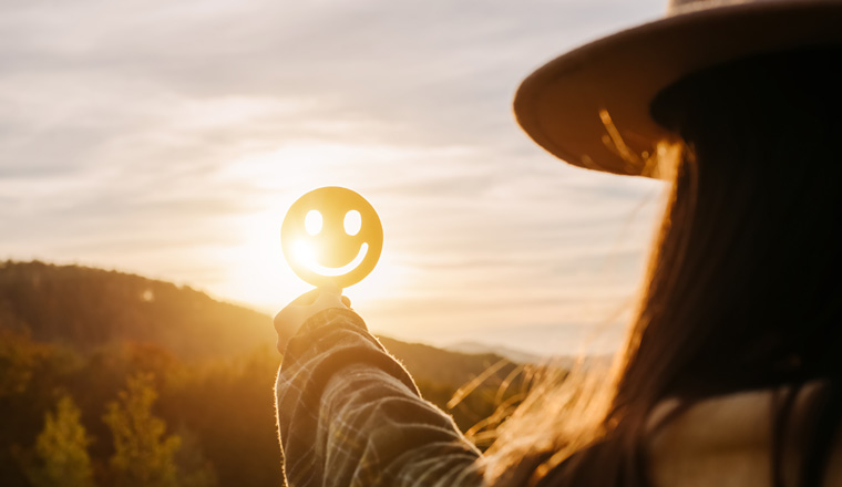 Close up of young woman holding happy smile face on background epic sunset in mountains at autumn sunny day, good feedback rating and positive customer review, satisfaction survey, mental health