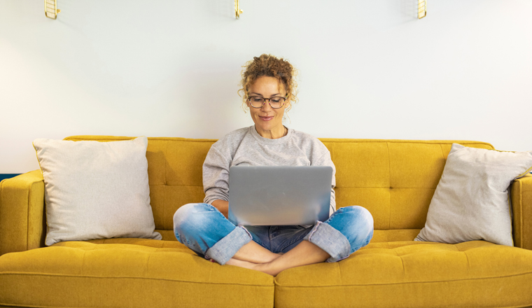 One woman smiling and using laptop computer at home sitting comfortably on a yellow sofa in living room. Smart working female people notebook. Surfing the web. Enjoying technology and connection