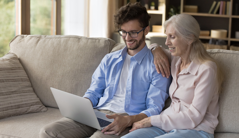 Positive grandson installing app on laptop, helping happy grandmother with software on computer. Cheerful young adult son and senior mom using pc for Internet online communication at home