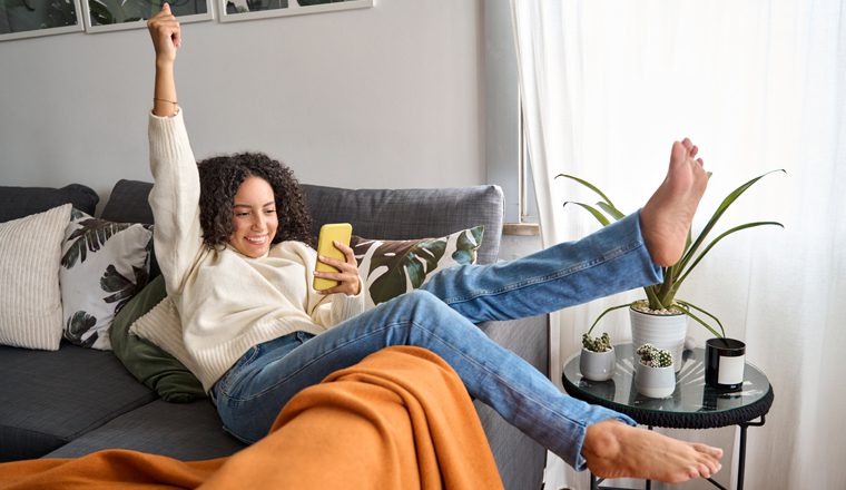 Happy excited young latin woman relaxing on couch using phone winning money in online app game. Young lucky woman feeling winner looking at cellphone, receiving great news or discount offer.
