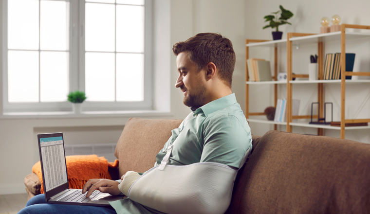 Businessman with broken arm working with laptop computer on his knees. Side view shot of recovery young man with bandaged arm sitting on sofa at home and working online