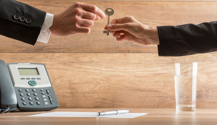 Businesswoman handing over a house key to a businessman in a conceptual image with their arms reaching across a desk with telephone and wooden background with copyspace.