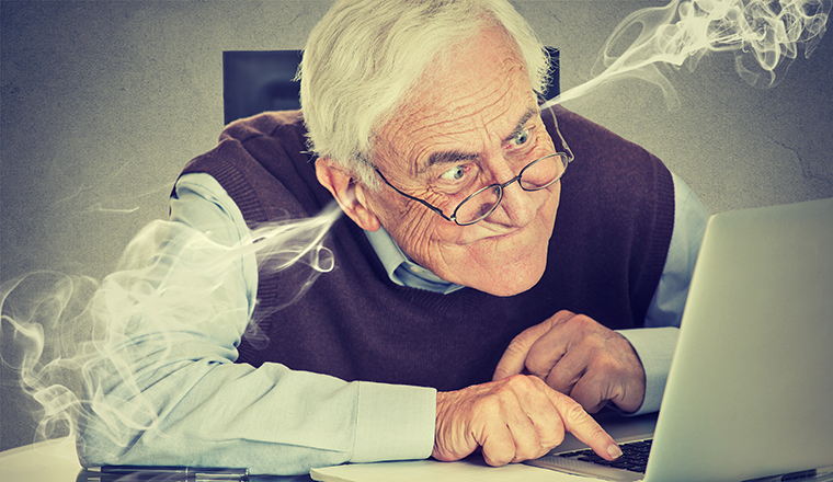 Stressed elderly old man using computer blowing steam from ears. Frustrated guy sitting at table working on laptop isolated on gray wall background. Senior people and technology concept 