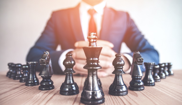Retro style image of a businessman with clasped hands planning strategy with chess figures on an old wooden table.