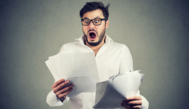 Young shocked guy in glasses looking at papers bank documents with list of bills and fees and feeling stressed on gray background