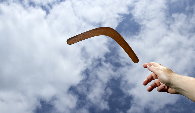 Throwing a plain wooden boomerang midair with blue sky and cloud background.