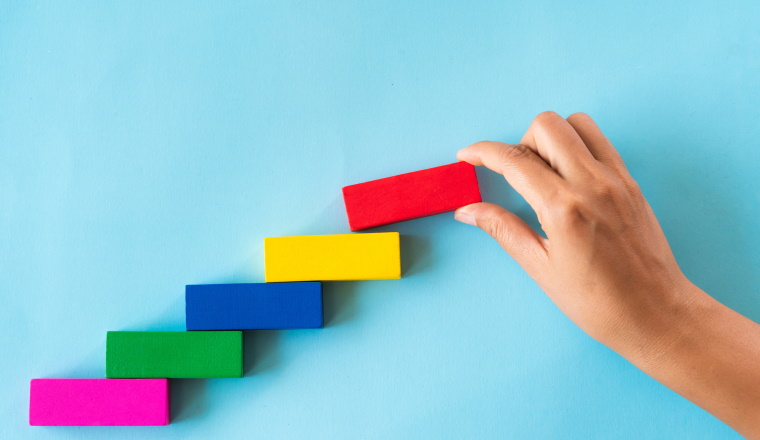 Concept of building success foundation. Women hand put red wooden block on colorful wooden blocks in the shape of a staircase