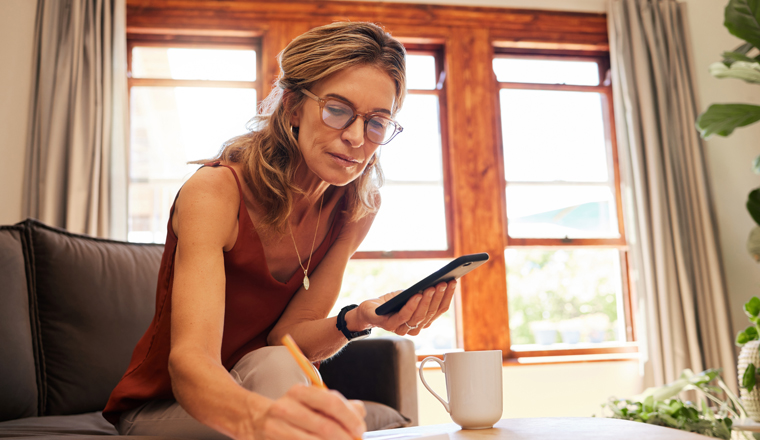 Phone, writing and senior woman planning retirement, taxes and budget on living room sofa of her house. Elderly person working on strategy for insurance in book with an email on mobile app with tech.