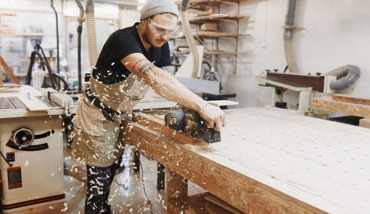 Carpenter working with electric planer on wooden plank in workshop.