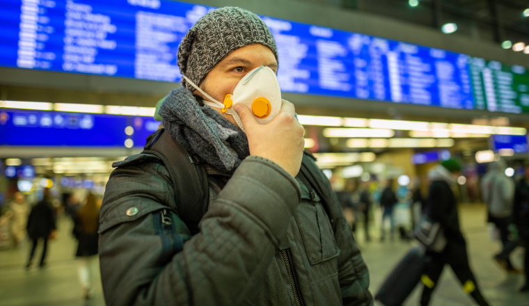 Man wearing a respirator mask device for health protection at an airport or railway train station in a crowd of people while travelling. Epidemic corona virus infection, flu sickness and travel illness concept.