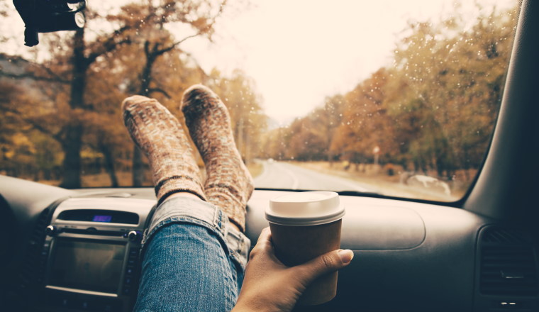 Woman feet in warm socks on car dashboard. Drinking take away coffee on road. Fall trip. Rain drops on windshield. Freedom travel concept. Autumn weekend. Filtered photo.
