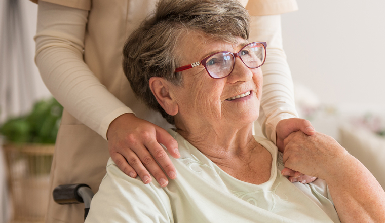 Panoramic view of elderly lady on wheelchair holding hands with supporting volunteer standing behind her