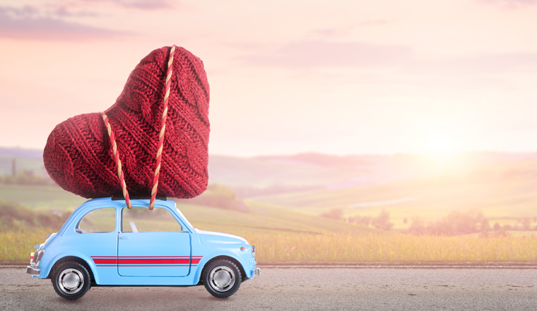 Blue retro toy car delivering heart for Valentine's day against blurred sunset rural landscape