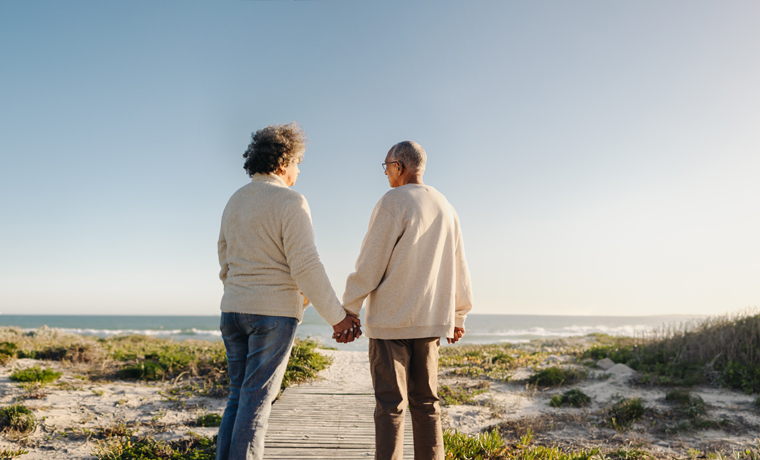 Panoramic view of a happy senior couple holding hands while walking down a foot bridge at the beach. Romantic elderly couple taking a refreshing seaside holiday after retirement.