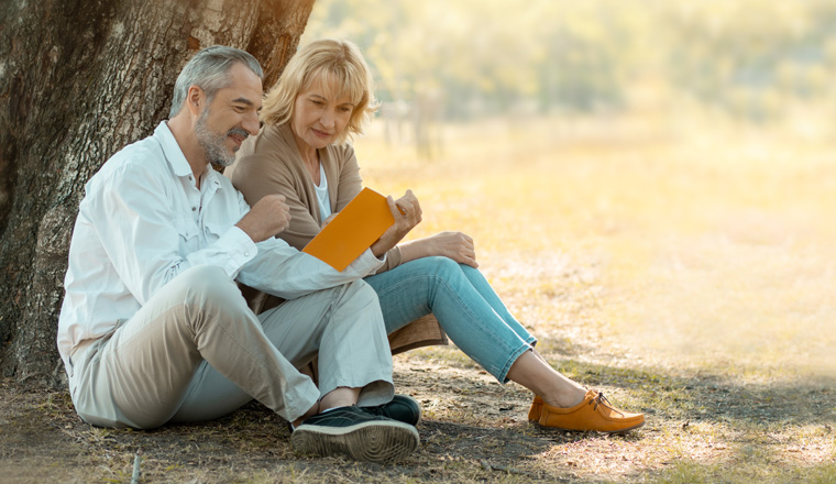 senior woman sitting on a fallen autumn leaves reading a book with husband or friend in a park, relax and vacation concept