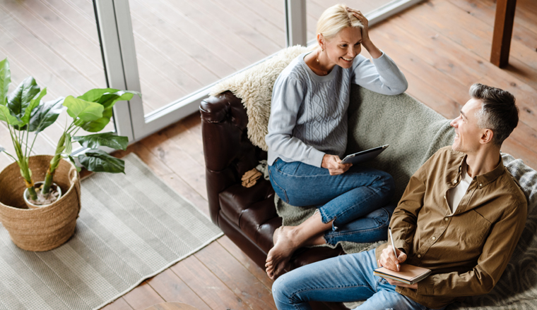 Smiling middle aged white couple relaxing on a couch at home, relaxing, using tablet, writing in notebook, top view