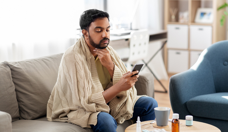 health, cold and people concept - sick young indian man in blanket with smartphone at home