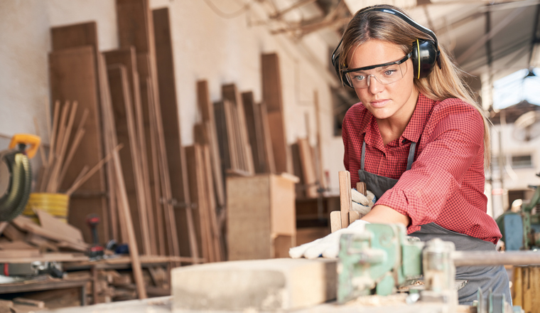 Woman as a carpenter trainee planing wood with a hand plane with hearing protection