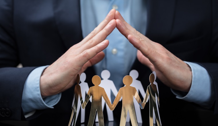 Close-up Of A Businessperson's Hand Protecting Paper Cut Out People On Desk