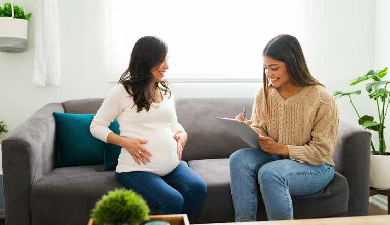 Smiling hispanic midwife writing information about the pregnancy and due date of a caucasian woman while sitting in the living room home