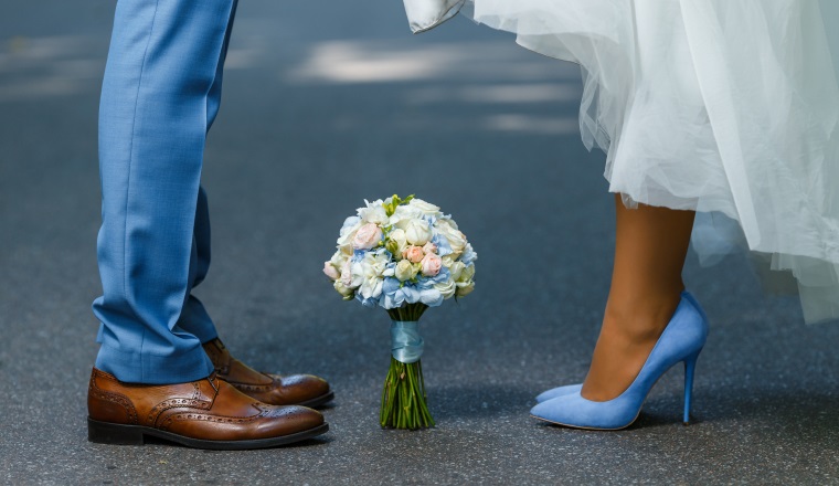 Wedding details: classic brown and blue shoes of bride and groom. Bouquet of roses standing on the ground between them. Newlyweds standing in front of each other