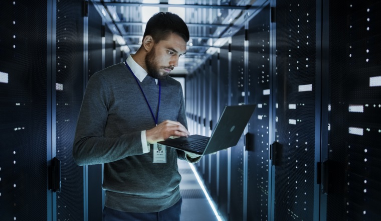 IT Technician Works on a Laptop in Big Data Center full of Rack Servers. He Runs Diagnostics and Maintenance, Sets up System.
