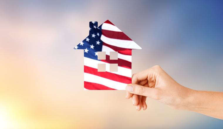 patriotism, home and citizenship concept - hand holding paper house in colors of american flag over evening sky background